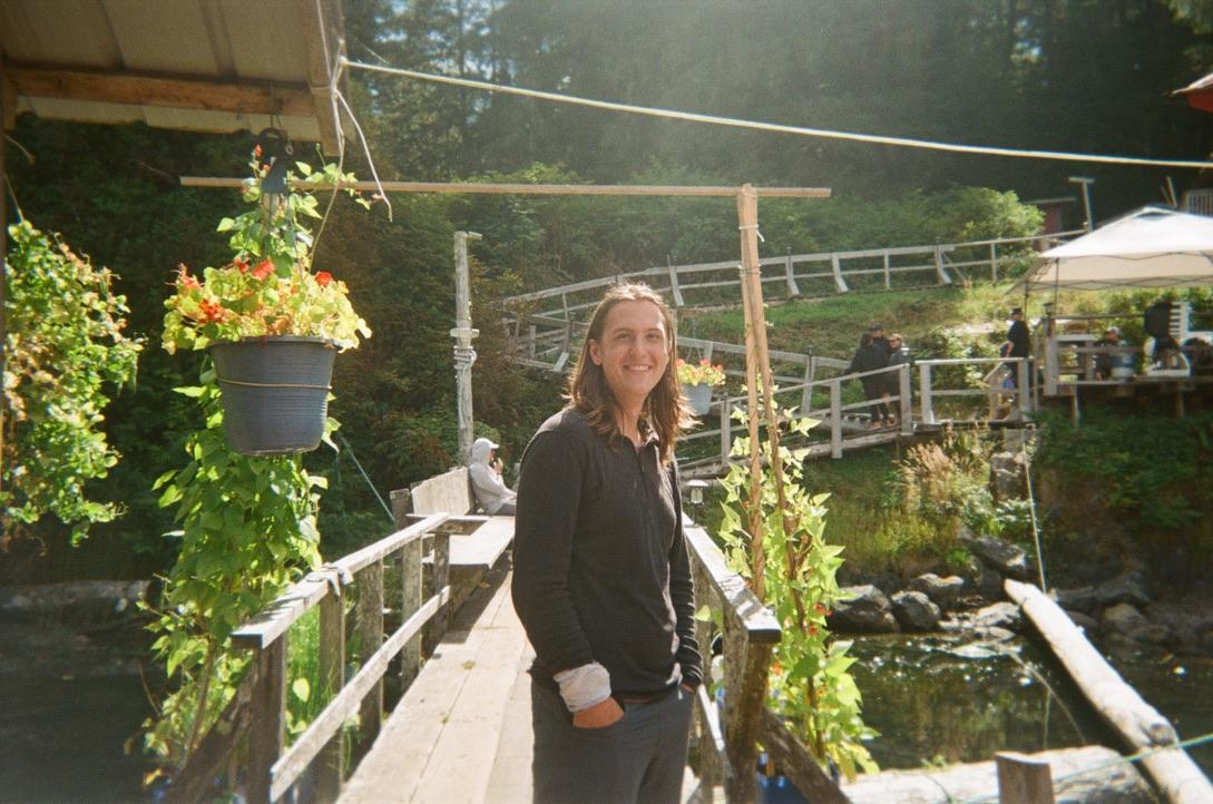 a photo of me, surrounded by plants in the sunlight, standing in front of a short bridge